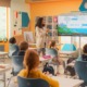 Curriculum fidelity k-12: Woman with long, dark hair in tan sweater and slacks stands at front of colorful classroom next to slide screen with a pointer explaining a colorful science slide to elementary students sitting at desks