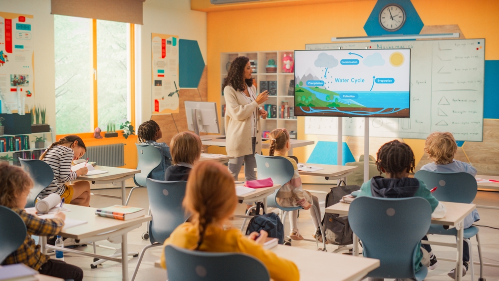 Curriculum fidelity k-12: Woman with long, dark hair in tan sweater and slacks stands at front of colorful classroom next to slide screen with a pointer explaining a colorful science slide to elementary students sitting at desks