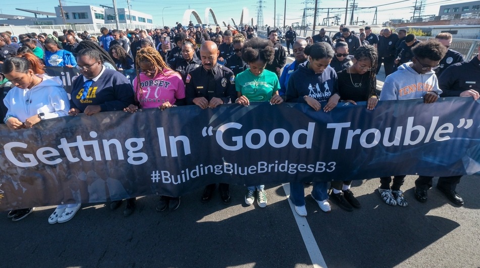 Police in schools: Large group of teens and adults holding dark blue banner with white text, "Getting in Good Trouble - Building Blue Bridges" stand in the front line of a street parade event.