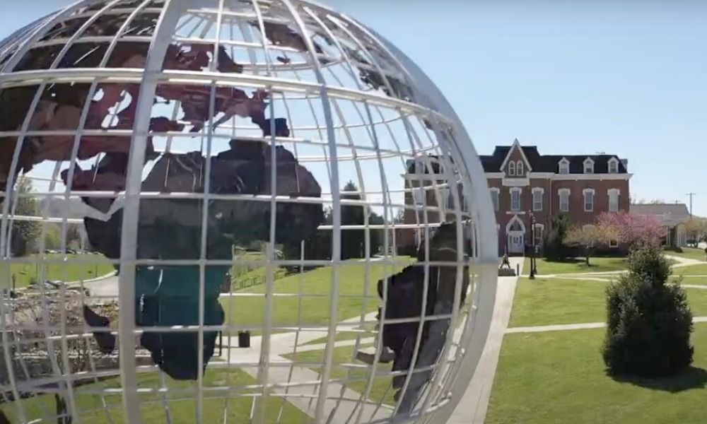 College listening cohort: Very large sculpture of white wireframe earth globe with blue, brown and green cutouts of continents suspended inside wire frame, with traditional multistory red brick building far in background after long green lawn