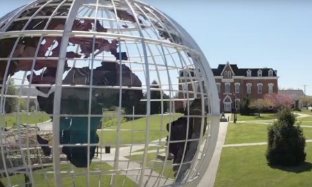 College listening cohort: Very large sculpture of white wireframe earth globe with blue, brown and green cutouts of continents suspended inside wire frame, with traditional multistory red brick building far in background after long green lawn