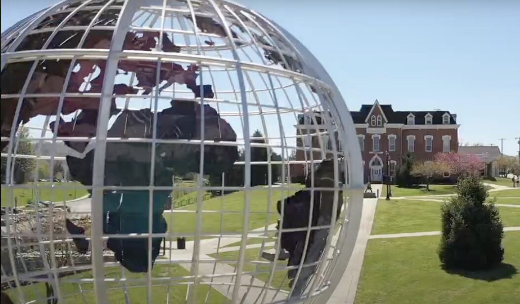 College listening cohort: Very large sculpture of white wireframe earth globe with blue, brown and green cutouts of continents suspended inside wire frame, with traditional multistory red brick building far in background after long green lawn