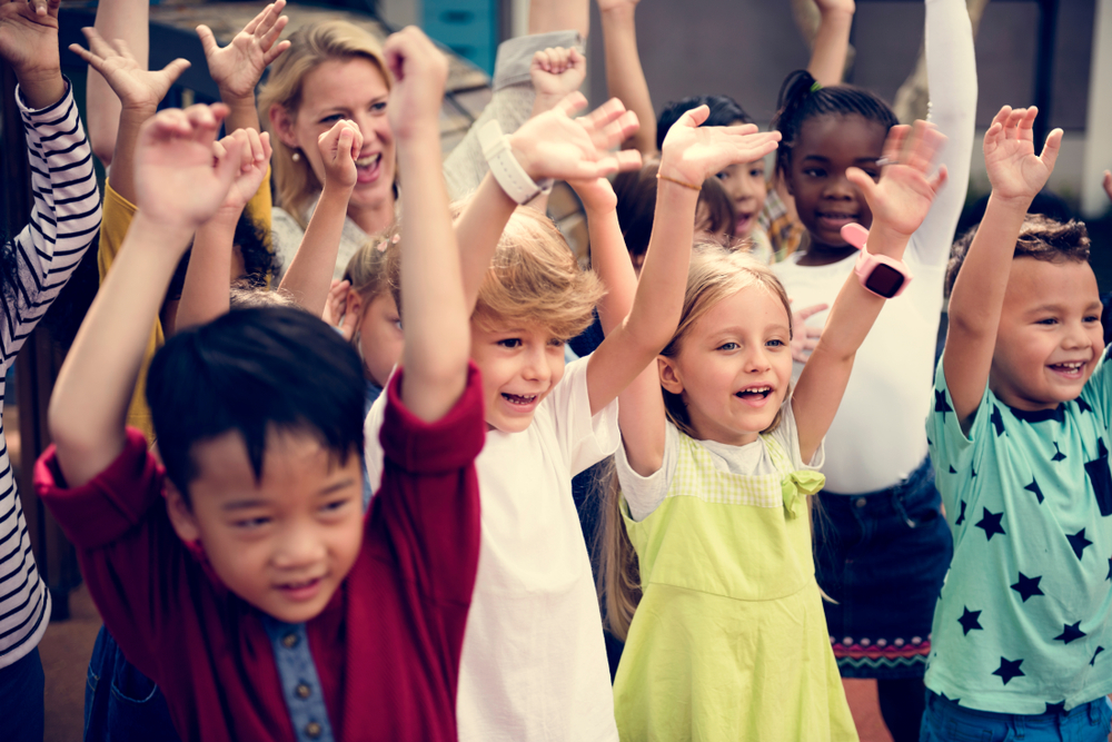 Childcare like Hunger Games: Several young children stand closely together with hands in air smiling into camera