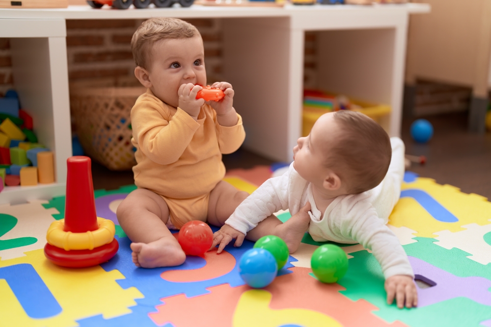 CHildcare like Hunger Games: Two young infants sit and lay on colorful floor ma playing with plastic toys