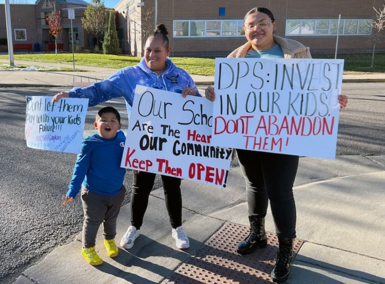 Education in 2025: 2 female adults and one young child stand outside an elementary school with hand-lettered protest signs about the school closing