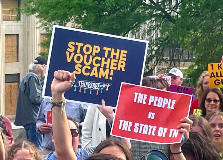 Education in 2025: Several adults stand outside with green trees in background holding navy and red protest signs about school vouchers