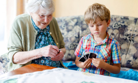 Intergenerational care: Senior womn and young boy sit next to each other on ouch both holding a and of cards and smiling while they look at their cards.