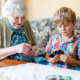 Intergenerational care: Senior womn and young boy sit next to each other on ouch both holding a and of cards and smiling while they look at their cards.