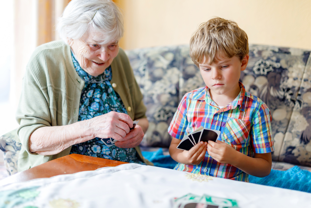 Intergenerational care: Senior womn and young boy sit next to each other on ouch both holding a and of cards and smiling while they look at their cards.