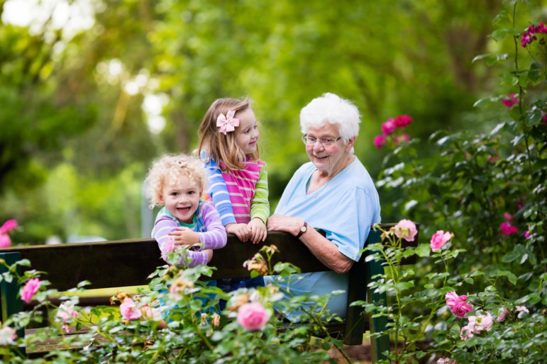 Intergenerational care: Older senior woman sits on bench with two young girls, all looking over back of bench into flower bed full of red and pink flowers