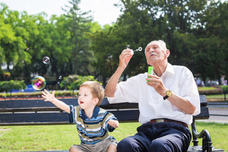 Intergenerational care: Older senior man sits on outdoor bench blowing bubbles next to toddler boy reaching out to cath a bubble