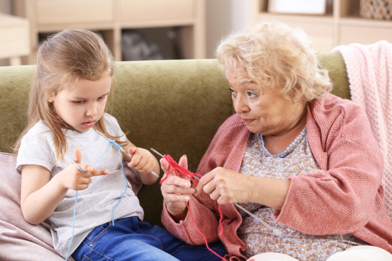 Intergenerational care: Senior woman with white hair shows a knitting stitch to young blonde girl trying to copy her, both sitting next to each other on a couch