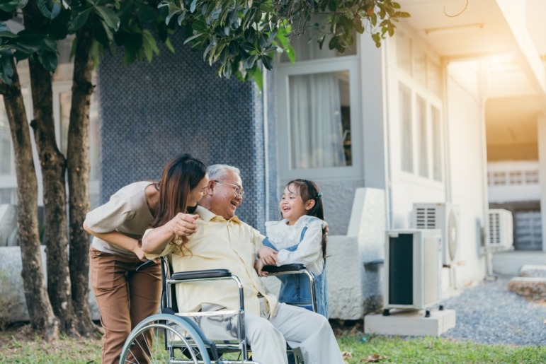 Intergenerational care:Young woman pushes wheelchair with elder senior man who smiles at young girl standing near chair smiling at him.