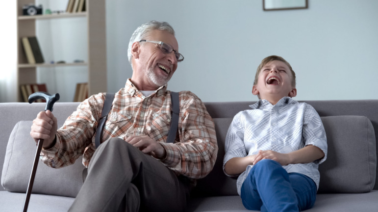 Intergenerational care: Older man holding cane and young boy sit on couch together laughing