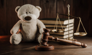 Migrant children: Tan teddy bear sits on dark tabletop next to stack of books, brass scales of justice and judges wooden mallet