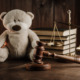 Migrant children: Tan teddy bear sits on dark tabletop next to stack of books, brass scales of justice and judges wooden mallet