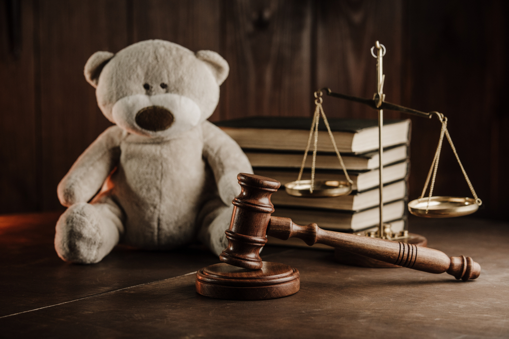 Migrant children: Tan teddy bear sits on dark tabletop next to stack of books, brass scales of justice and judges wooden mallet