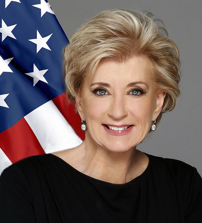 9 education topics: Headshot blonde woman with short hair in black top in front of American flag