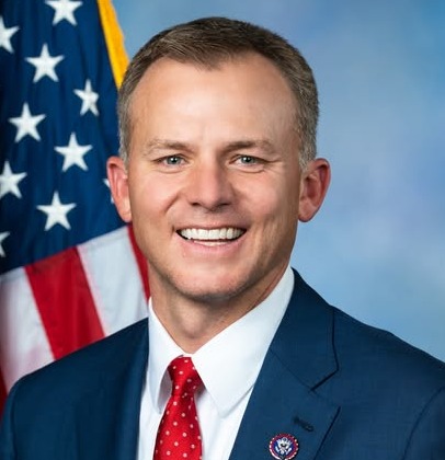 9 education topics: Headshot man with short light brown hair in navy suit, white shirt and red tie in front of American Flag