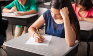 National test scores low: Closeup of girl with long, dark hair, with head resting on fist, sitting at desk taking a test. A few students sitting behind her are visible.