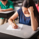 National test scores low: Closeup of girl with long, dark hair, with head resting on fist, sitting at desk taking a test. A few students sitting behind her are visible.