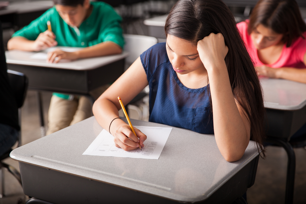 National test scores low: Closeup of girl with long, dark hair, with head resting on fist, sitting at desk taking a test. A few students sitting behind her are visible.