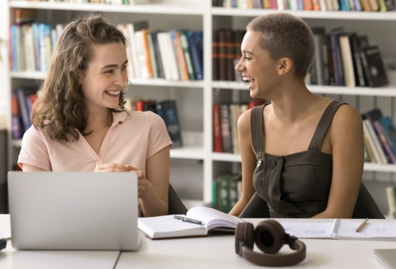 Generational empathy: Two older teen girls sit at desk with laptops looking at each other and smiling