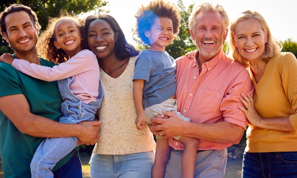 Generational empathy: 2 senior adults, 2 adults in their thirties, and two children being held by adults, stand in a line smiling into camera.