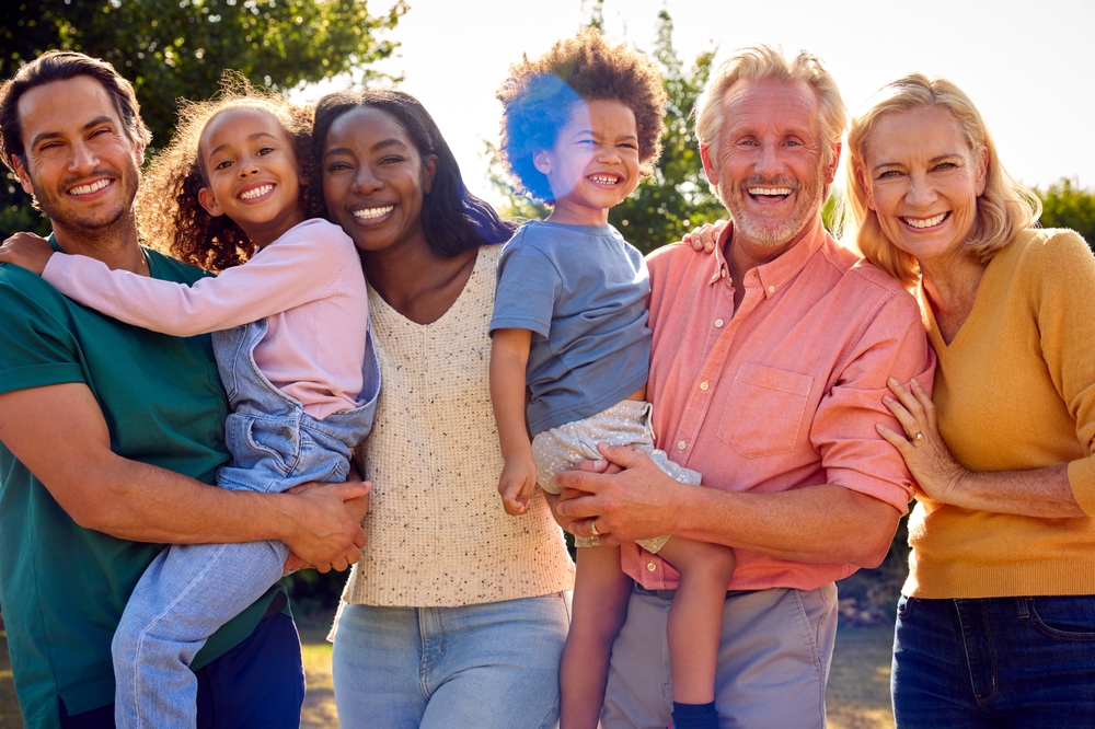 Generational empathy: 2 senior adults, 2 adults in their thirties, and two children being held by adults, stand in a line smiling into camera.