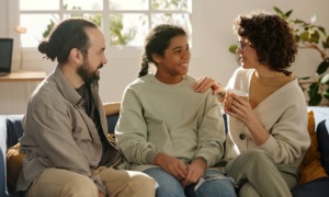 Parenting challenged kids: Two dark-haired adults - one man & one woman - sit on a couch with a dark-haired female teen between them. All three are smiling and looking at each other smiling.