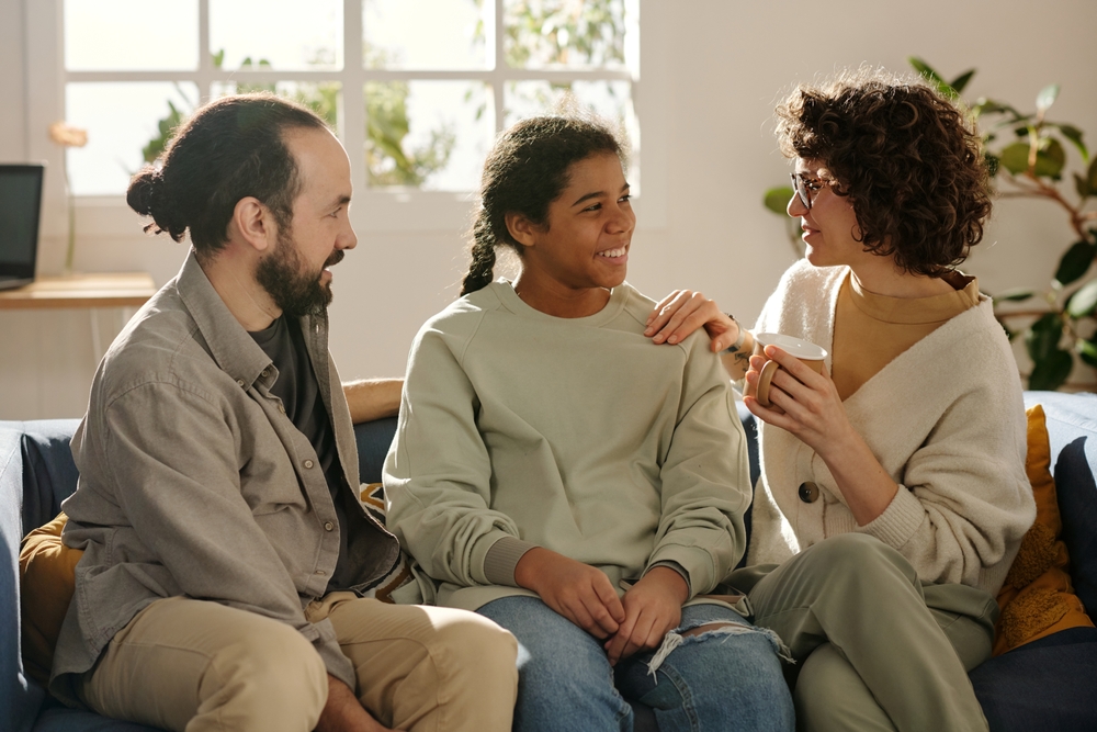 Parenting challenged kids: Two dark-haired adults - one man & one woman - sit on a couch with a dark-haired female teen between them. All three are smiling and looking at each other smiling.
