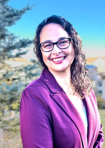 Katherine Plog Martinez: white woman with long, dark curly hair, black rim glasses, wearing purple jacket, smiling outside in front of trees
