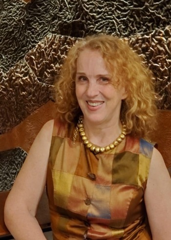 Merita Irby headshot: Woman with blonde and red. curly hair wearing sleeveless gold print dress smiles into camera