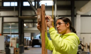 Union high school apprenticeships: Older teen girl with dark hair puled back in bright yellow jacket holds hands up working with tool on pieces of wood above her head