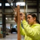 Union high school apprenticeships: Older teen girl with dark hair puled back in bright yellow jacket holds hands up working with tool on pieces of wood above her head