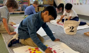 Math building blocks: Several kindergarten students sit on gray carpeted floor working with colored chips and numbered white cards