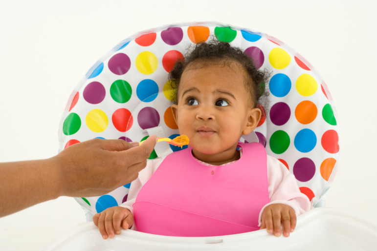 QR code baby food heavy metal data: Infant in igh chair with brigh colored polka dots on cushion back with adult hand in foreground bringing spoonful of baby food to baby's mouth