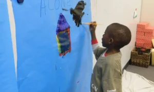 Childhood grief: Ypung Black boy stands painting in black and red on a very large sky blue piece of paper tacked to wall