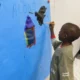 Childhood grief: Ypung Black boy stands painting in black and red on a very large sky blue piece of paper tacked to wall