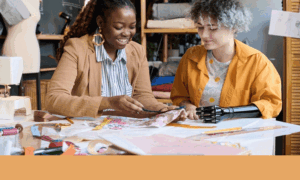 Disabled student programs DOGE cuts: Teen girl with black artificial arm sits with adult Black woman at table with sewing machines pinning cloth to pattern pieces