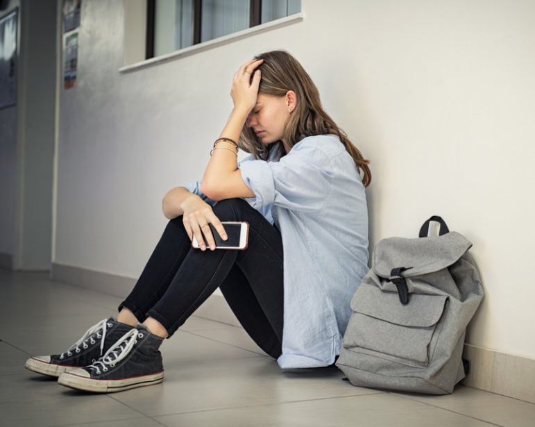 LGBT teen girl suicide: Older teen with long light brown hair, wearing black leggings, over-sized light blue shirt and black high top shoes, sits on sidewalk with next to gray backpack leaning against white building with knees up and head in hand