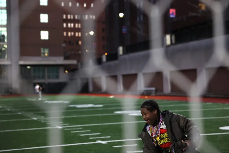Disabled student job training: Young Black teen boy in brown winter jacket and t-shirt with brightly colored red and yellow graphic seen through chainlink fence carrying football and running around green soccer field with multi-storied red brick buildings in background