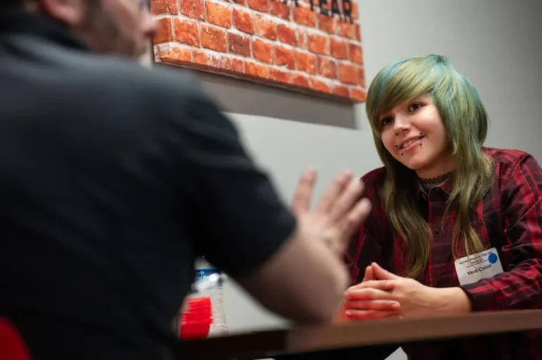 Disabled student job training: Closeup of man' in black polo shirt sitting across table from young female teen with blue streaked blonde hair in a red plaid shirt