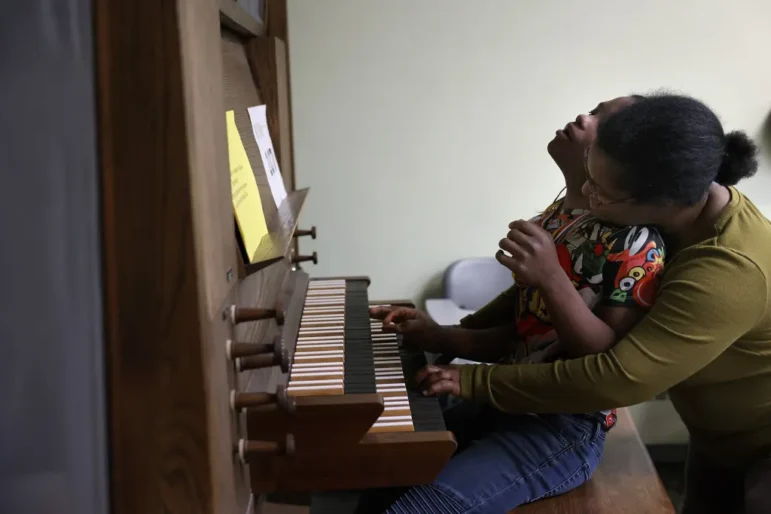 Disabled student job training: Young bBlack male teen sits at piano with Black woman standing behind him helping him place his hands on the keys