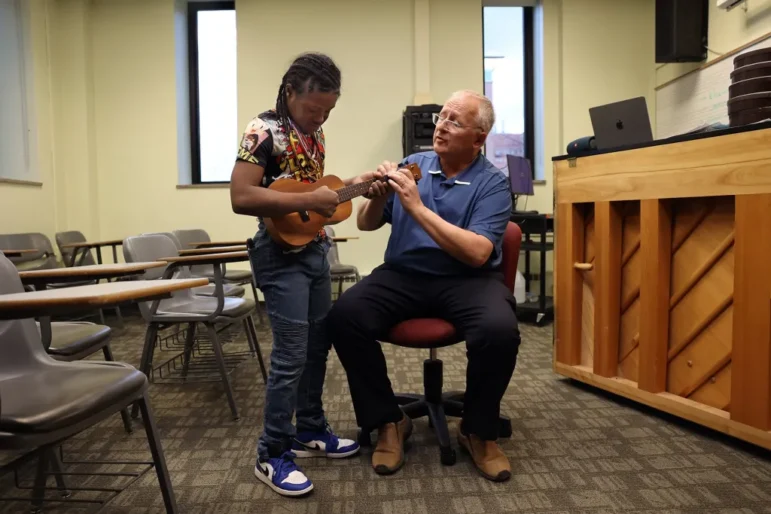 Disabled student job training: Young Black boy in red print shirt and blue jeans stands plays ukulele as older man with gray hair in blue shirt and black pants places hands and fingers on strings at neck of ukulele instructing him on how to play
