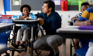 Disabled education from DOE to HHS: Young Black boy in wheelchair sits at desk in elementary classroom with biracial male adult crouched on floor next to him speaking to him about classwork