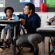 Disabled education from DOE to HHS: Young Black boy in wheelchair sits at desk in elementary classroom with biracial male adult crouched on floor next to him speaking to him about classwork