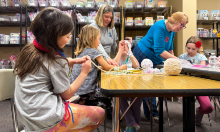 Cell phone alternative activities: Several elementary-age students and two elder women sit around a large table knitting.
