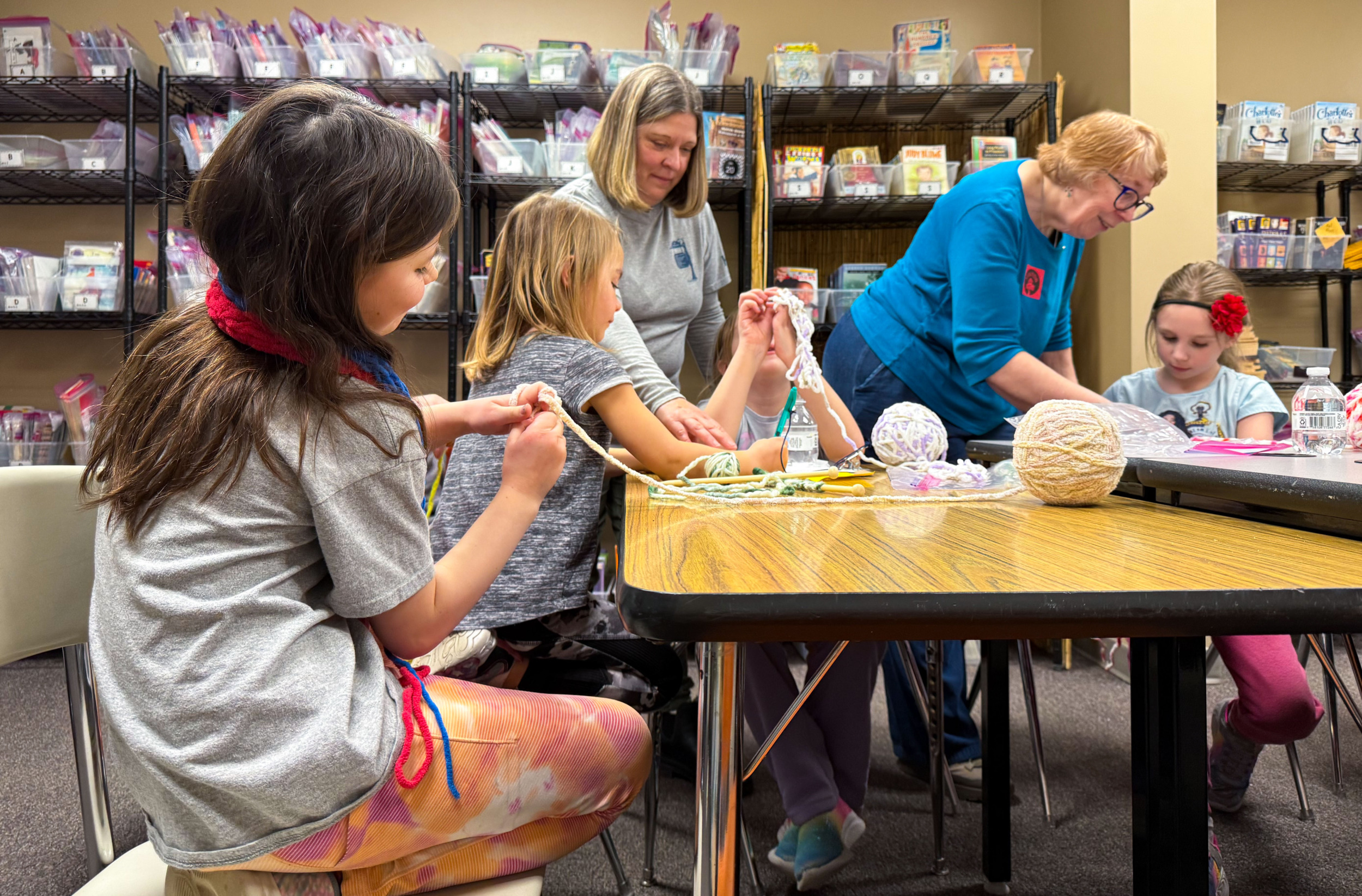 Cell phone alternative activities: Several elementary-age students and two elder women sit around a large table knitting.