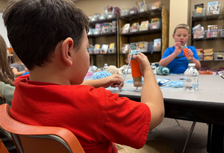 Cell phone alternative activities: Elementary-age boy with drk hair in red shrt sits working with yarn at table